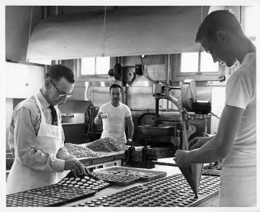 Black-and-white photo of workers preparing pastries in the Miller & Paine bakery, with trays of dough lined in rows on a worktable and large industrial equipment in the background.