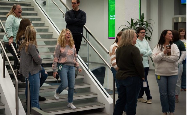Group of people gathered inside Nelnet Center near a staircase during a fundraising event, with several individuals standing and watching an activity.