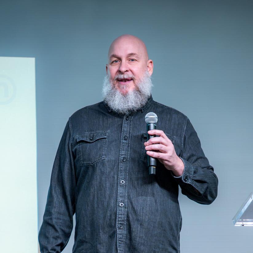 Photograph of a presenter speaking into a microphone during a presentation. A screen beside the presenter displays Green Squirrel branding concepts and merchandise examples, while an audience member is visible in the foreground.